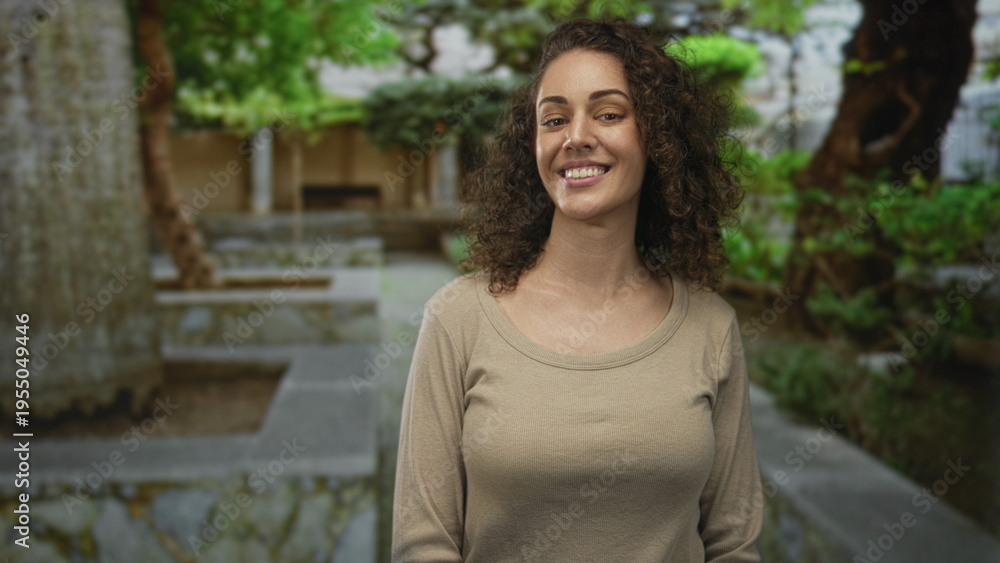 Fototapeta premium Woman smiling at camera with curly hair and visible neck, standing on a stone park walkway beside a low stone building and trees, wearing a beige sweater; warmth connection happiness.