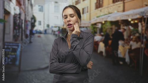 Woman holding cheek with hand, grimacing from toothache at a street cafe with arms crossed; oral pain distress.