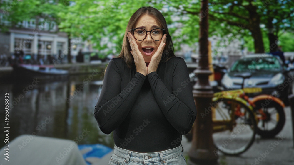 Fototapeta premium Young woman touching hands to cheeks in surprise on street beside a canal with parked bicycle and cars; surprise wonder.