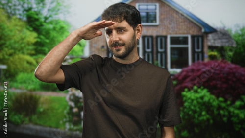 Man with beard wearing brown tshirt saluting with hand to forehead in front of building garden pathway; respect contemplative.