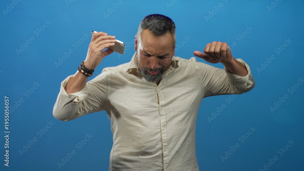 Fototapeta premium Middle age man with beard holding a smartphone and raising his arm while dancing against a bright blue studio backdrop; joy celebration.