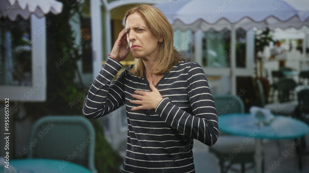 Fototapeta premium Woman touching ear then clutching chest in street cafe near outdoor tables and umbrellas, gingerly raising hands toward throat with pained expression; distress.