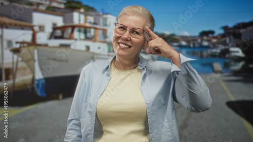 Fotografia Woman points finger to head on a seaside street with boats and dock visible; joyful travel memory recollection