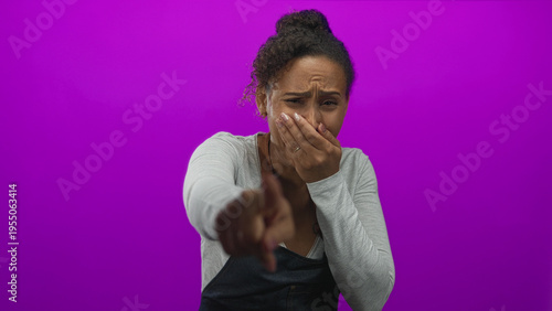 Canvas Print Woman pointing finger at camera and covering her mouth in a purple studio backdrop wearing a denim apron and bun hairstyle; embarrassment teasing