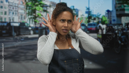 Fényképezés Woman cupping ear with hand and furrowed brow on a busy city street, wearing denim apron and long sleeve top; concern listening