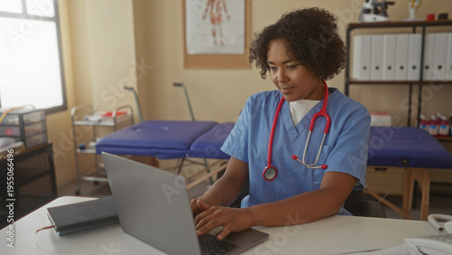 Wallpaper Mural Young african american woman in blue scrubs typing on laptop at white desk in clinic office; serenity productivity. Torontodigital.ca