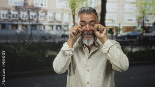 Fotografie Man with grey beard rubbing eyes and raising hands in a startled gesture on an amsterdam street by a canal and buildings; surprise tiredness