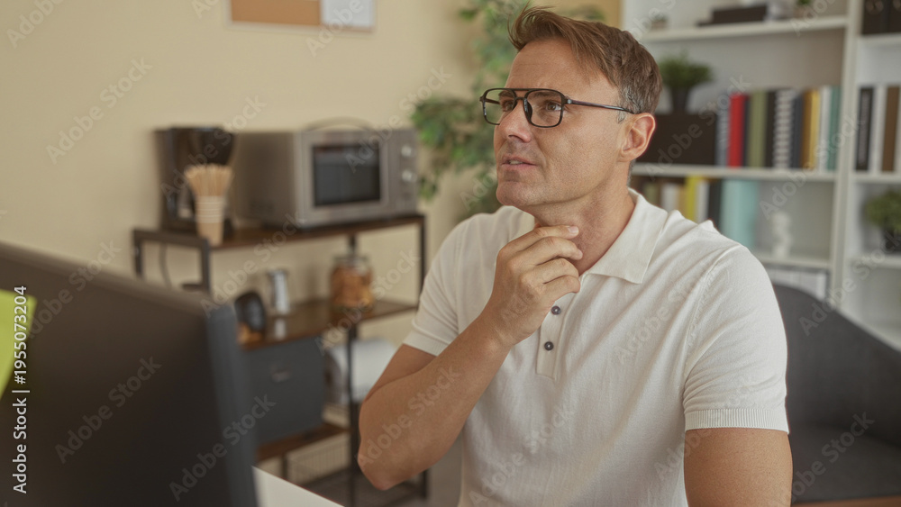 Fototapeta premium Man wearing glasses and white polo touching chin while looking at a computer screen in an office building at his desk; thoughtful focus.