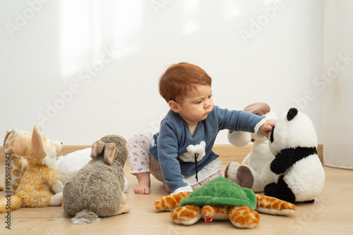 9-month-old baby girl sitting on the floor surrounded by plush toys and reaching for a panda plush with curiosity. 