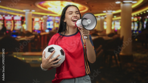 Obraz na plátne Woman in red jersey holding soccer ball shouts into megaphone inside building with casino slot machines visible; excitement