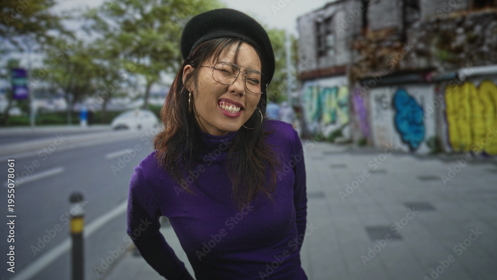 Fototapeta premium Woman wearing beret and glasses tilts head with eyes closed and hands behind back on city street beside graffiti wall, purple turtleneck visible; playful ease.