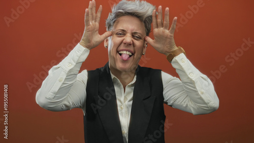 Bild auf Leinwand Woman framing her face with both hands and sticking out her tongue in a studio with an orange wall; playfulness