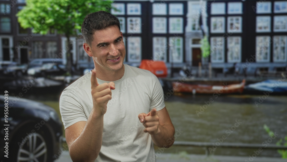 Fototapeta premium Man points finger with both hands on a canal street in amsterdam near boats and cars, smiling in a white t shirt; playful confidence.