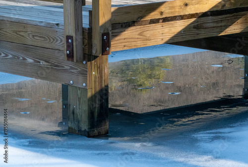 Schilderij op canvas Sunlit Wooden Dock Structure with Strong Shadows Reflected in Partially Frozen W