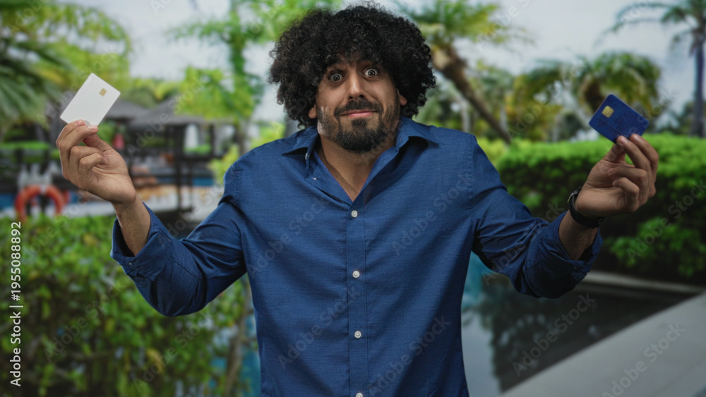 Fototapeta premium Man holding two creditcards in both hands at a building pool area showing a hesitant shrug; financial uncertainty.