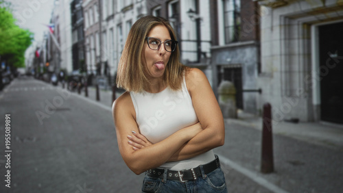 Фотография Woman with arms crossed and tongue out on a city street beside bollards and stone buildings, wearing white tank top and glasses; playful defiance