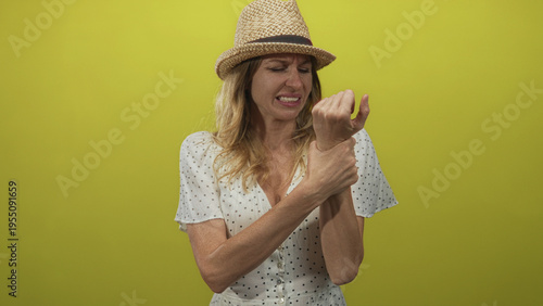 Foto Woman holds wrist in yellow studio wearing straw hat and polka dot dress, grimacing in discomfort; pain concern discomfort