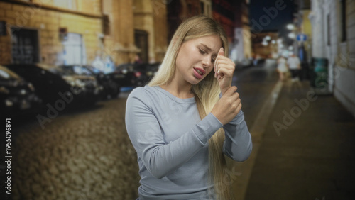 Fotografía Woman holding her wrist with a pained grimace on a dim cobblestone street at night, wearing a light sweater; pain concern unease