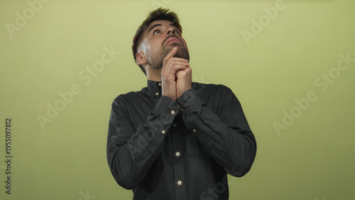 Bild auf Leinwand Young hesitant hispanic man buttoning shirt collar with both hands against light green wall in studio; fear anxiety dread worry