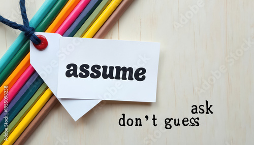 White tag with assume written on it near colorful pencils. Text ask dont guess encourages inquiry not speculation. Background is light wood. Reminder to seek information.