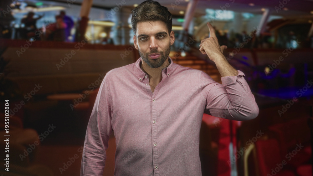 Fototapeta premium Man with beard beckoning with hand in airport lounge, wearing pink shirt and standing near seats and railing; assertion confidence.