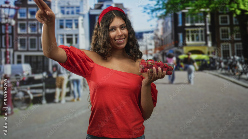 Obraz premium Hispanic teenage woman holding strawberries in hand on street market, red off shoulder blouse and headband visible; joy summer freshness.