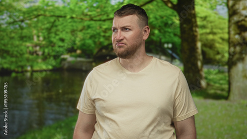 Foto Man grimacing with visible face and stubble, wearing beige tshirt, standing in forest by a pond and small bridge among trees and grass; disgust