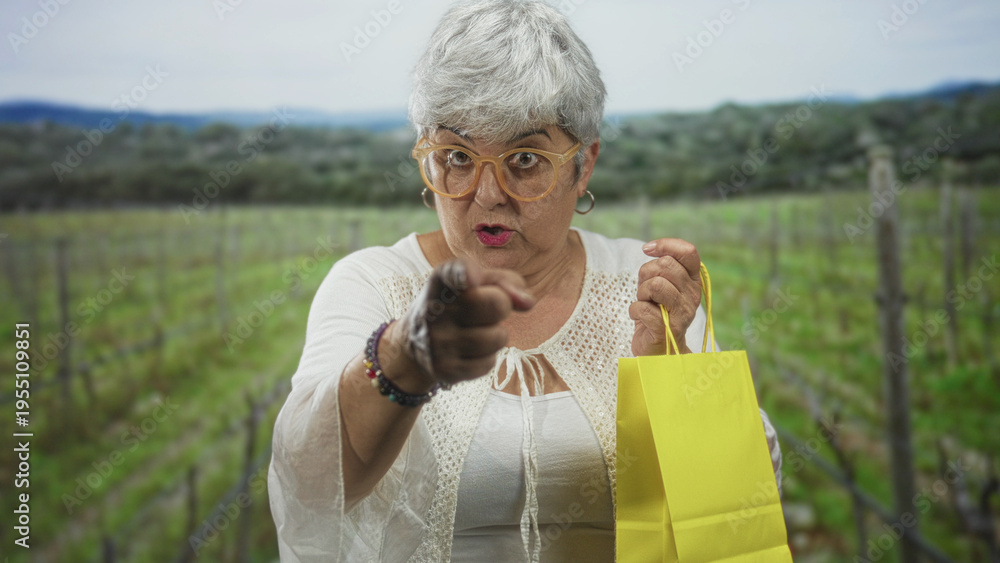 Obraz premium Senior woman with gray hair and glasses points finger while holding a yellow shopping bag in a vineyard among vine rows; assertive invitation.