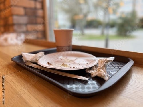Used disposable plates cup on tray closeup, crumpled napkins and crumbs on plastic tray by window on wooden counter, takeaway