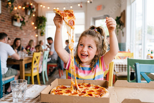 Cheerful young girl pulling a cheesy slice of pepperoni pizza in a busy family restaurant