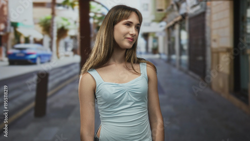 Young woman smiling and walking in a light blue tanktop on a narrow city street, looking sideways toward storefronts; carefree joy. Free Download Young woman smiling and walking in a light blue tanktop on a narrow city street, looking sideways toward storefronts; carefree joy. - GFXLoad