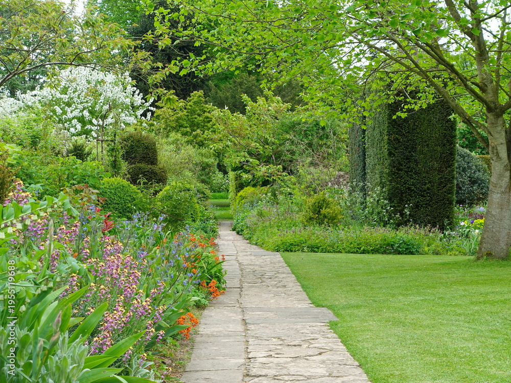 Naklejka premium A stone paved path passes through vibrant flower borders, lush grass lawn and manicured hedges. This formal garden landscape showcases professional landscaping and horticultural excellence.