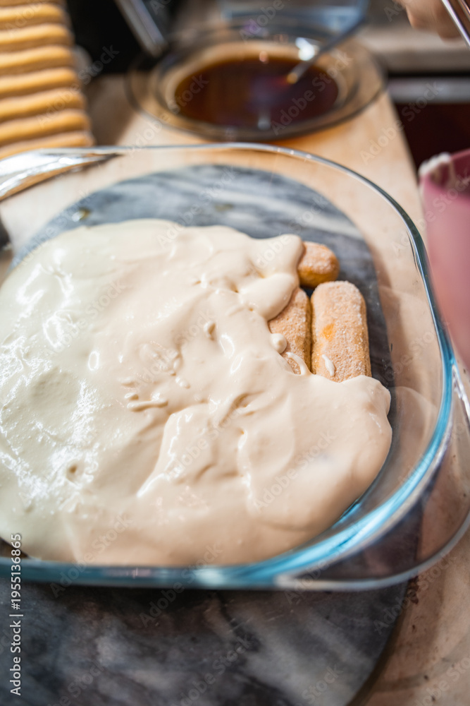 Fototapeta premium Homemade Tiramisu preparation in a glass baking dish, layering creamy mascarpone over espresso-soaked ladyfingers, a traditional Italian dessert in progress.