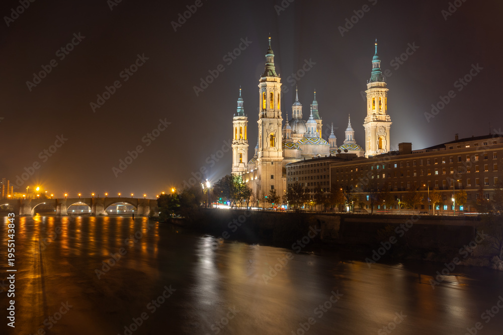 Fototapeta premium Night cityscape of Basilica del Pilar with light trails and reflections on the Ebro river in Zaragoza, Spain. Illuminated historic architecture and urban skyline.