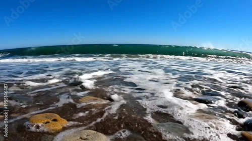 Low angle view of ocean waves crashing onto a pebbled beach on a bright sunny day against a clear blue sky