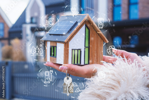 Woman holding model house with solar panels and keys with digital eco icons overlay. Concept of sustainable real estate, smart home technology, renewable energy savings and green housing investment.