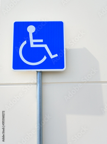 A blue and white sign showing the symbol for accessible parking is attached to a pole. It is positioned on a nearby building wall in a city. The sun casts a shadow on the wall