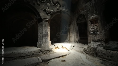 Walking through a dark doorway into the ancient rock-cut Geghard Monastery in Armenia. A shaft of light illuminates intricate stone carvings before revealing a dim room with candles.

