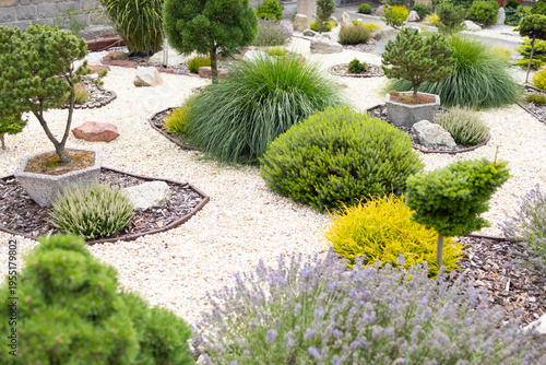 Lush garden landscape featuring various ornamental shrubs, grasses, and stones arranged in a decorative pattern with gravel pathways and vibrant greenery