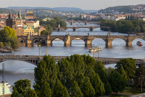 Scenic view of multiple historic bridges spanning the Vltava River in Prague, with lush greenery and city architecture visible along the riverbanks