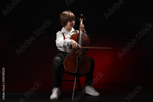 Schilderij op canvas Little boy playing violoncello on dark red background