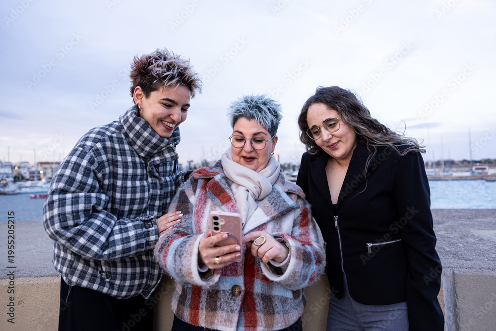 Obraz premium Mother and daughter with a friend using a smartphone outdoors by the sea. Family, friendship and digital connection in a relaxed coastal urban environment.