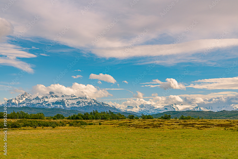 Obraz premium The iconic snow-covered peaks of the Paine Massif rise above green grasslands under blue sky with scattered clouds at Torres Del Paine National Park, Chilean Patagonia.