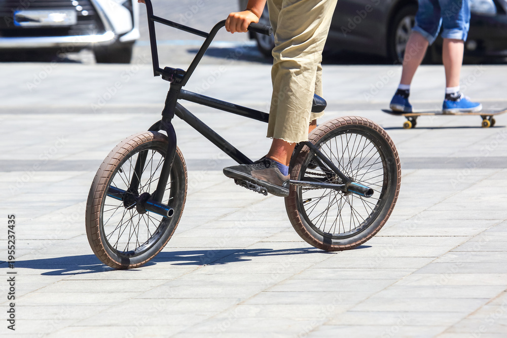 Fototapeta premium Biker rides a black BMX bike on pavement while a person skateboards in the background during a sunny day