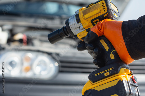 Fotografi Close up of a mechanic hand wearing an orange safety glove holding a professional battery powered impact driver