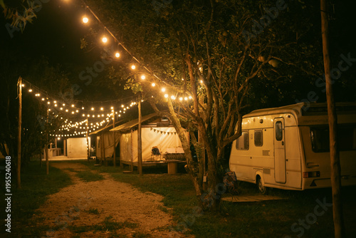 Camp party outdoor holiday relax scene with glowing string lights above caravan and tent in forest night atmosphere perfect for joyful gathering and peaceful escape