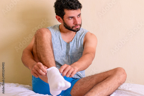 Photos Handsome man putting adjusting socks while sitting on the bed, looking to the side, resting