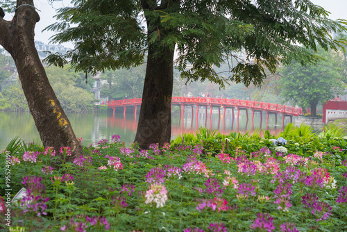 A peaceful garden scene with blooming flowers and large trees overlooking Hoan Kiem Lake and its red bridge during misty pre Tet days in Hanoi.
