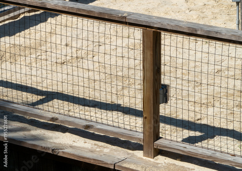 Фотография Weathered wooden railing and wire mesh fence beside a sandy path, casting long shadows under bright daylight