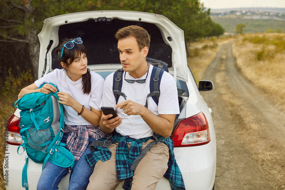 Obraz premium Couple by car use phone, map, backpack. Stranded on a rural road, travelers check geolocation, seek signal, and study a map while resting on trunk near forest. Adventure on weekend travel and hiking.